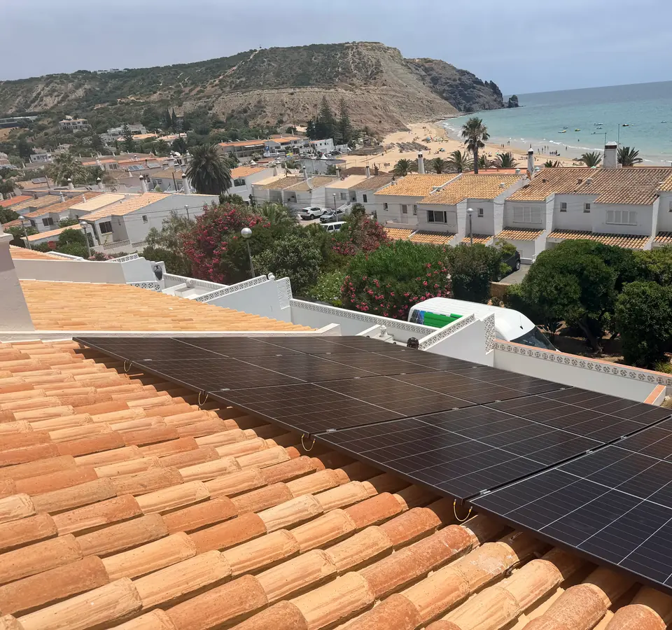 Painéis solares escuros no telhado de uma moradia na Praia da Luz, com a baía e a praia ao fundo.